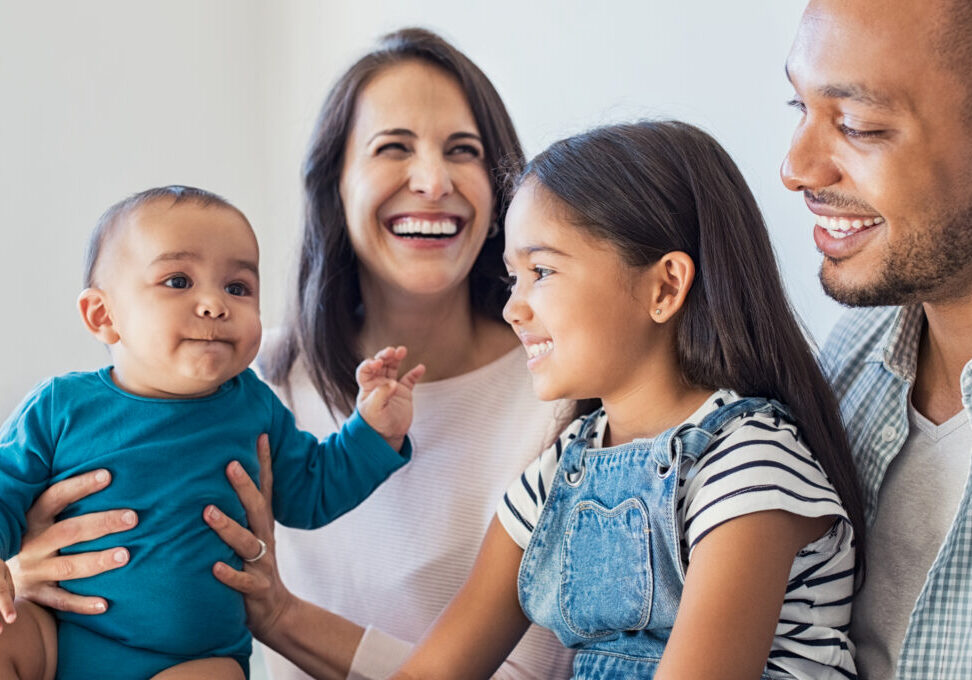 Happy daughter sitting on father legs and looking his little brother. Excited infant playing with elder sister. Portrait of smiling multiethnic family sit on sfoa and enjoying at home.