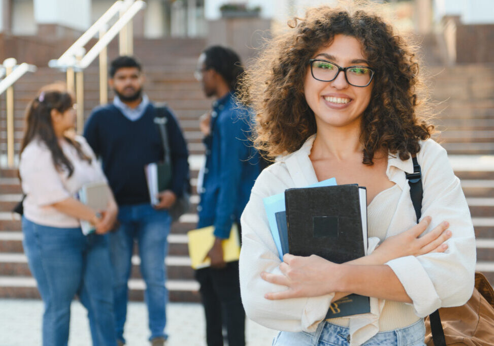 Happy young woman student with books smiling on university campus, diverse students standing in background