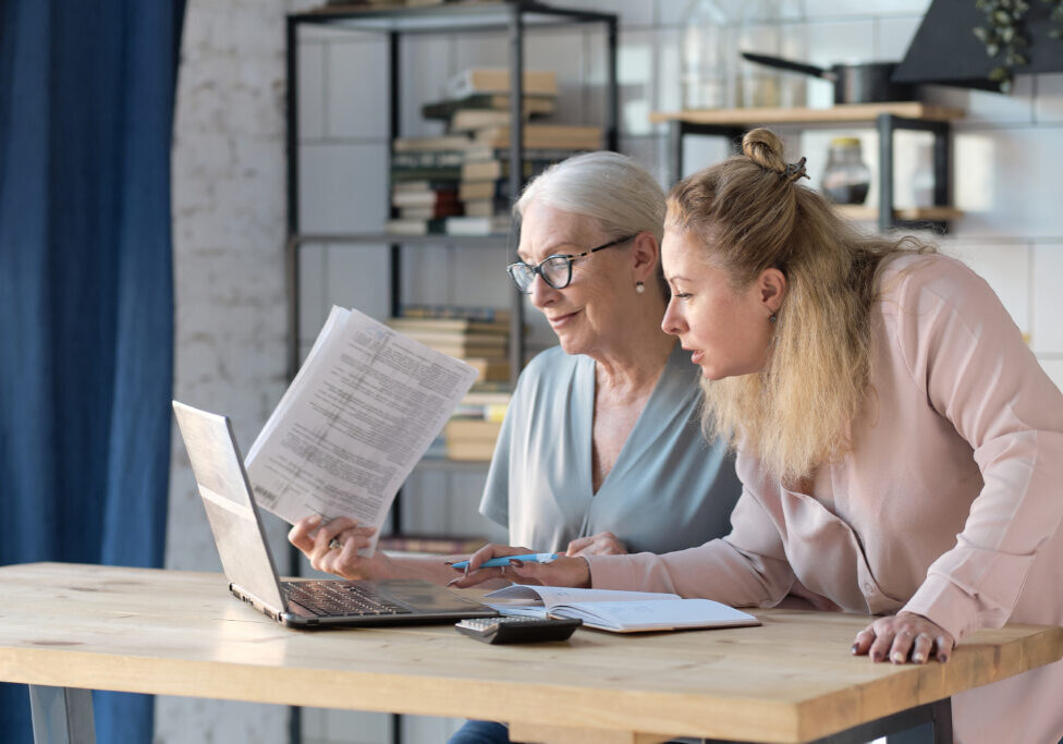 Senior woman using laptop for websurfing in her kitchen. middle-aged daughter helps her mother with documents. Mature lady sitting at work typing a notebook computer in an home office