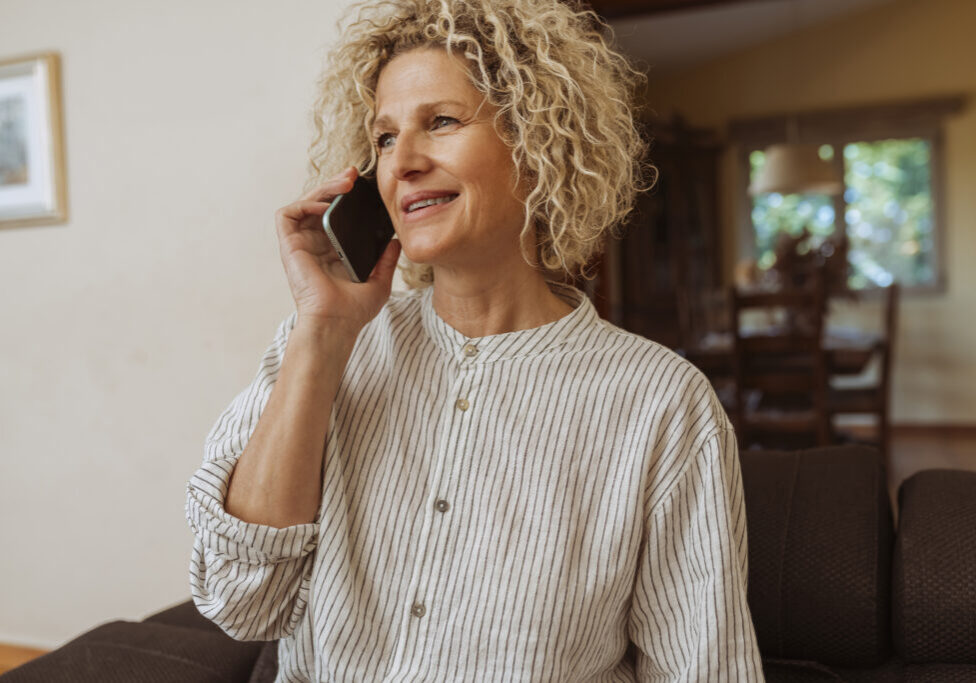 Happy middle aged woman using smartphone, enjoying pleasant conversation while relaxing on sofa in living room.