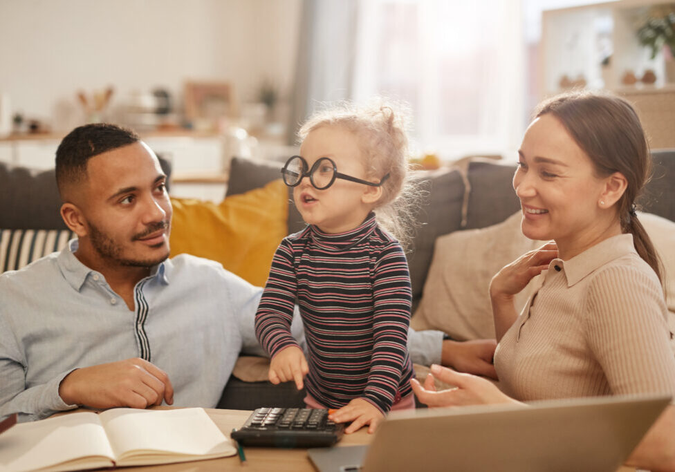 Portrait of happy young family counting home finances with cute little girl wearing glasses in sunlit apartment